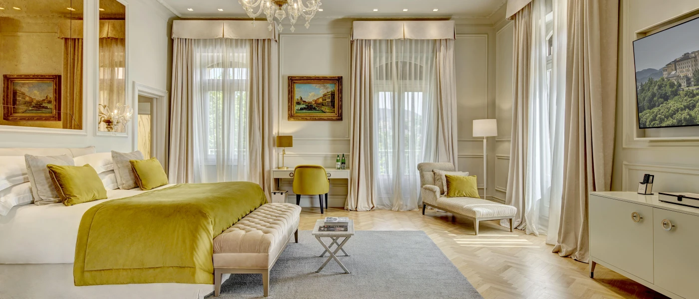 Hotel bedroom with high ceilings, gold soft furnishings, a chandelier and large windows dressed in sheer and beige curtains