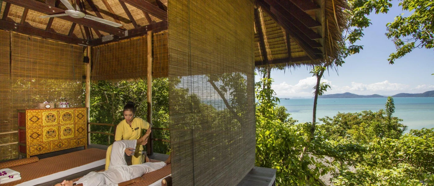Woman in white lies on a bed in a thatched open-air treatment cabana, as a staff member in yellow lifts her leg, with the ocean in the background