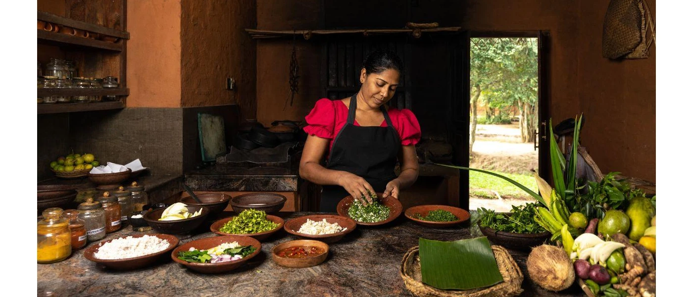 Woman in a black apron and pink blouse using her hands to mix herbs in a bowl, with bowls of food laid out in front of her