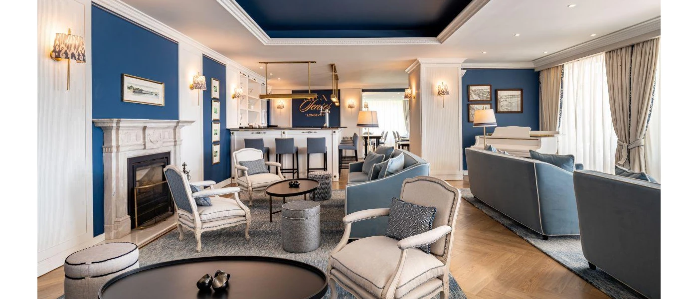 Cosy lounge with blue walls and sofas, a grey rug, gold lighting and white chairs with grey/blue cushions