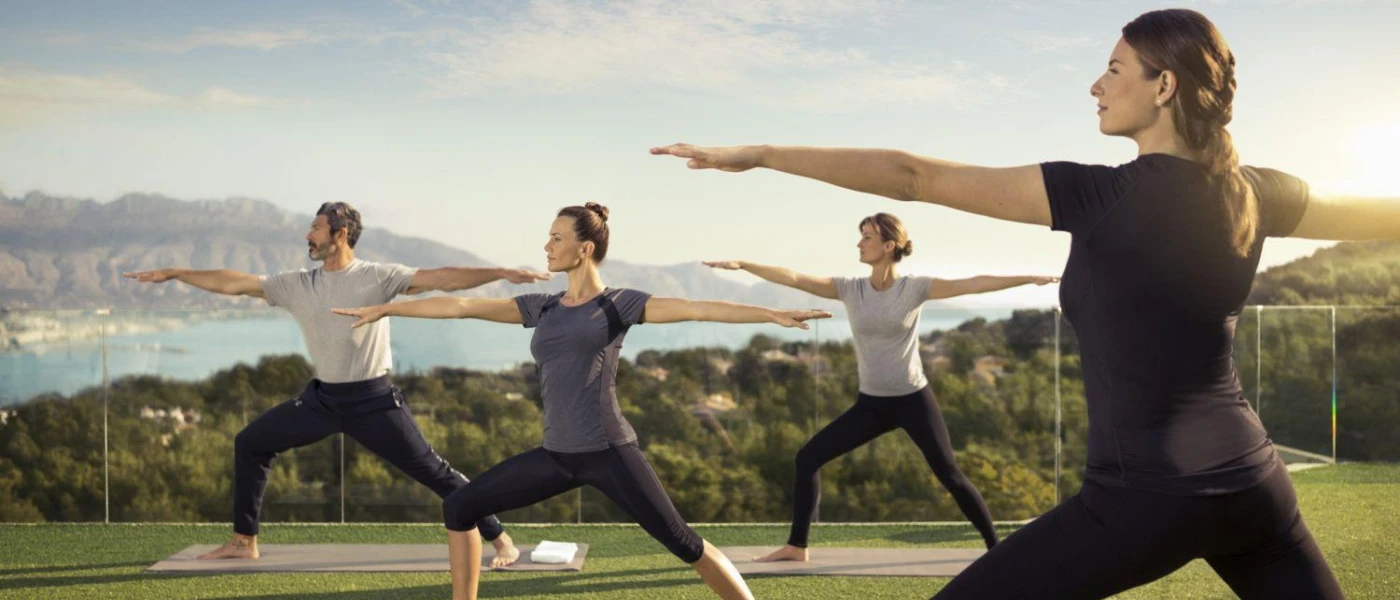 Group stand in warrior pose during a yoga session in a garden overlooking mountains