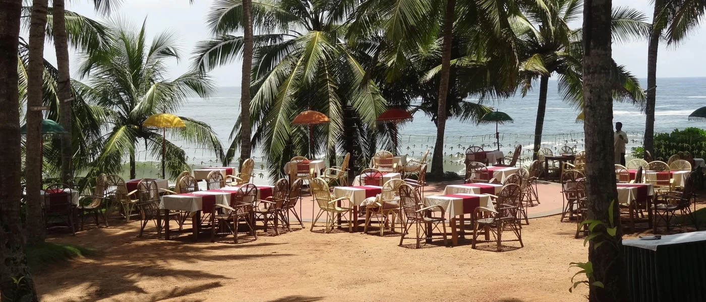 Wooden tables and chairs set up ready for guests to dine al fresco on the sand, surrounded by palms and metres from the ocean
