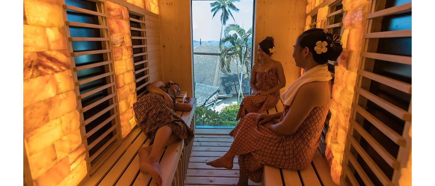 Three smiling women with flowers in their hair wearing patterned robes chat in a softly lit wooden sauna
