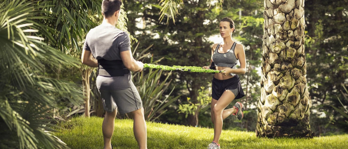 Man and woman in grey active wear work out using an elasticated rope in a lush garden setting