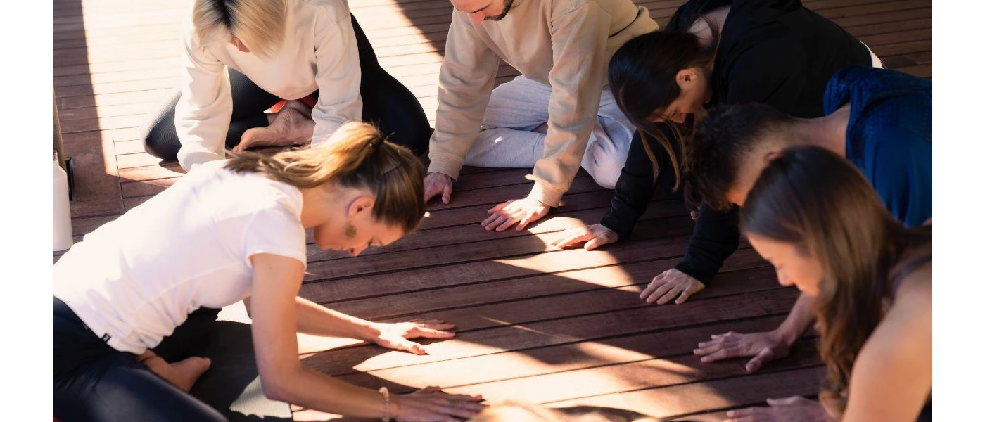 Group in comfy clothes sit in a circle on a wooden deck, with a person in the centre and their heads bowed