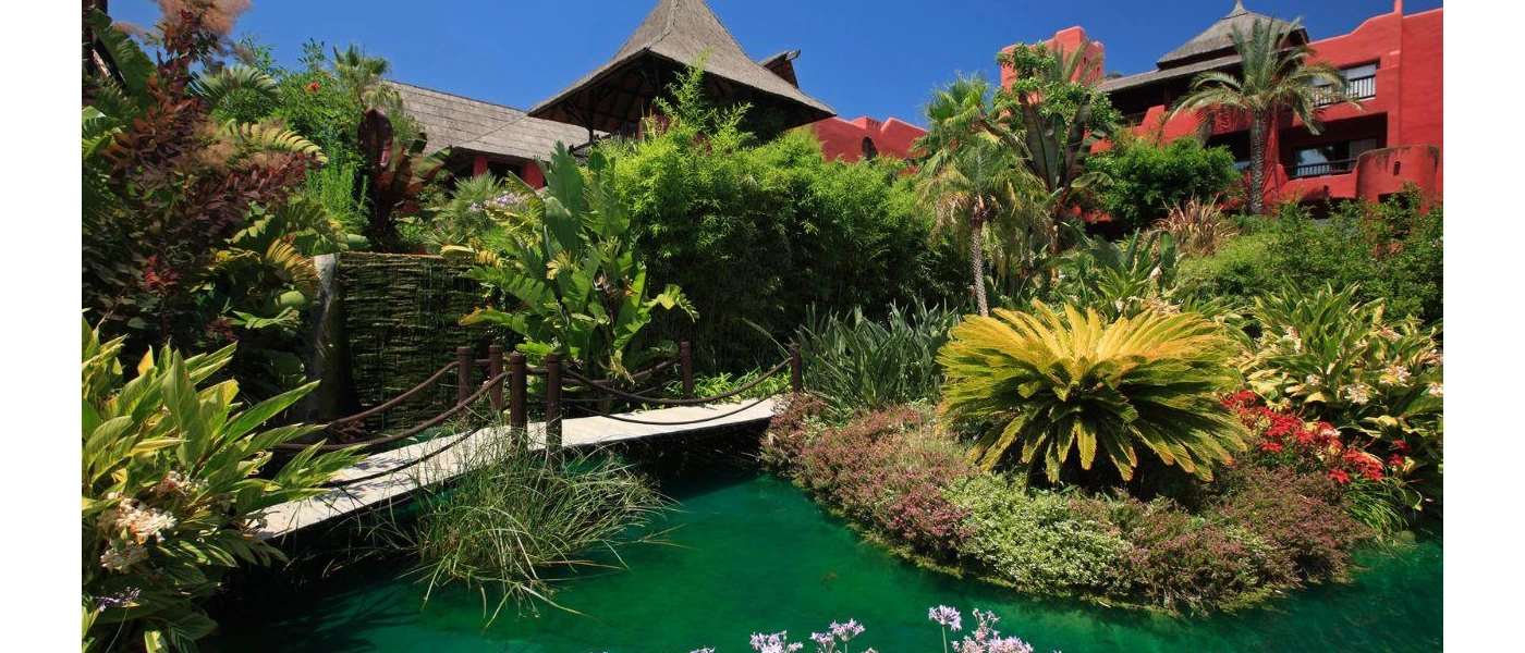 Tropical gardens with a pond and a roped footbridge next to red buildings with a thatched roof
