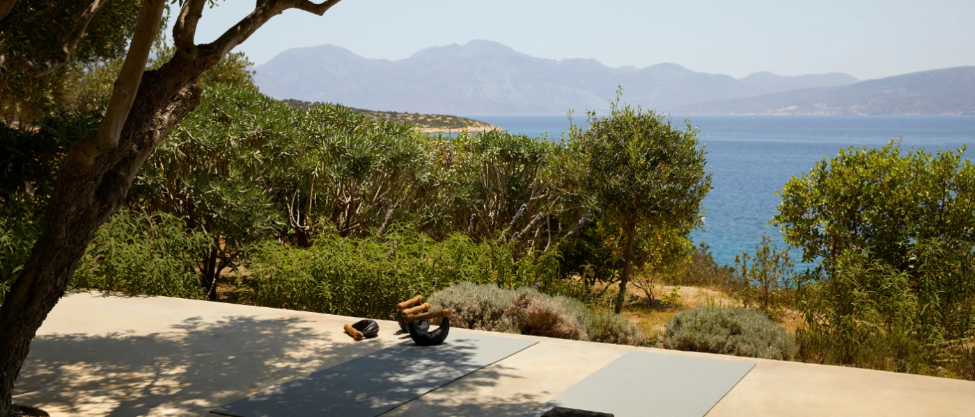 Grey yoga mats lined up on a stone terrace overlooking a green hillside and the sea