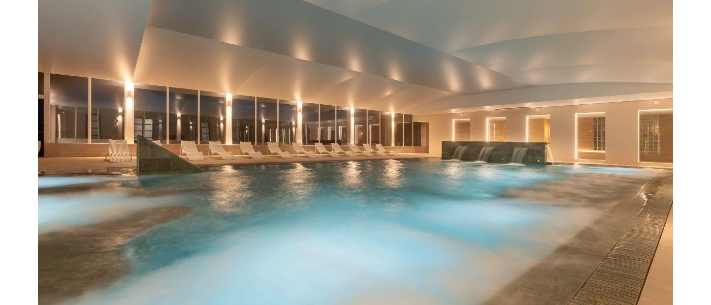 Indoor swimming pool at night-time, as seen through surrounding windows, with LED lighting and water jets