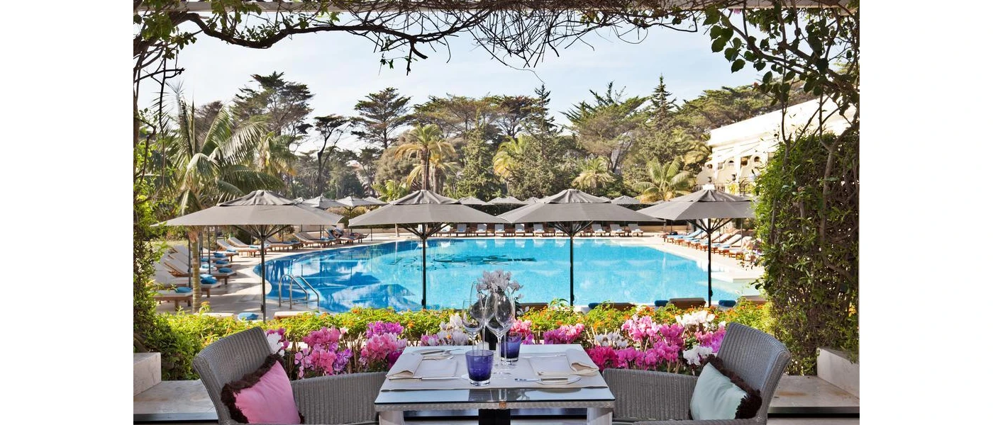 Outdoor swimming pool surrounded by grey parasols and white-cushioned loungers, tropical gardens and a terrace set up for dining with a bed of pink flowers overlooking the pool