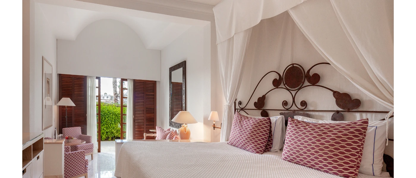 Chic hotel bedroom with white bedding and drapes, black iron headboard, pink patterned cushions and armchairs and wooden shutters opening onto a terrace fringed by a bush
