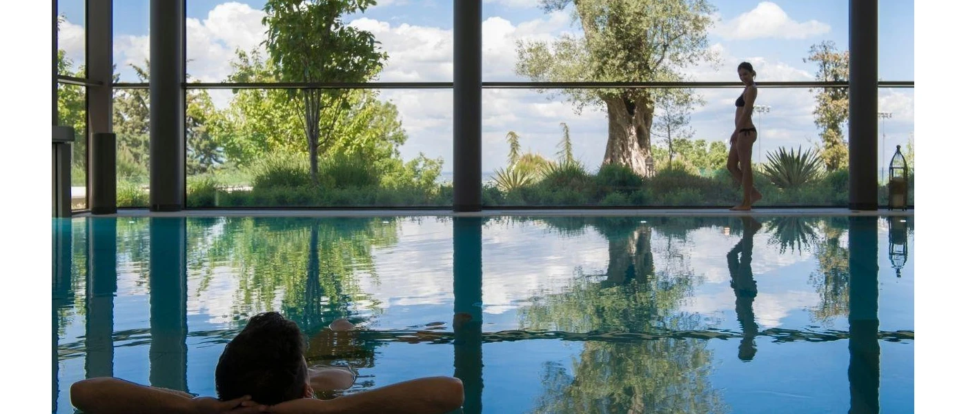 Man relaxing with his hands behind his head in an indoor swimming pool with floor-to-ceiling windows and views of outdoor greenery