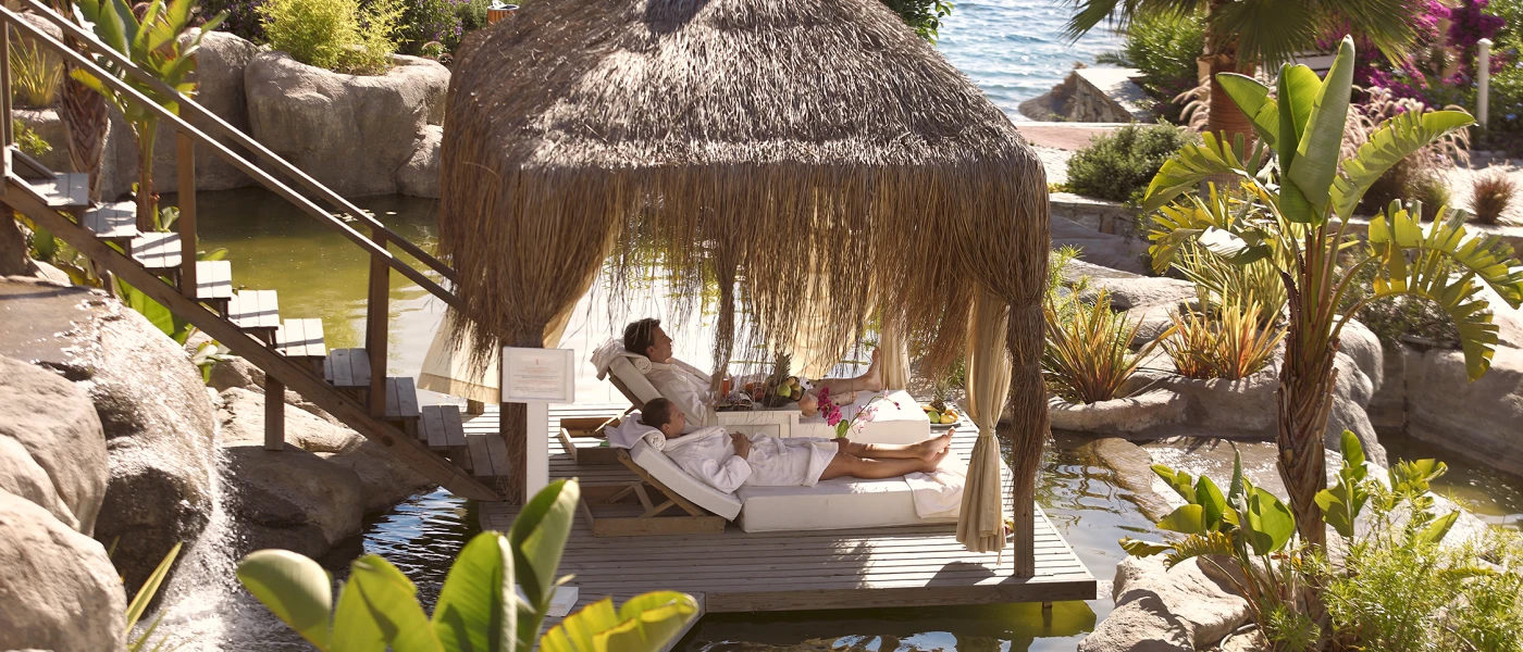 Couple in white robes relax on loungers in a thatched-roof cabana surrounded ponds and tropical gardens