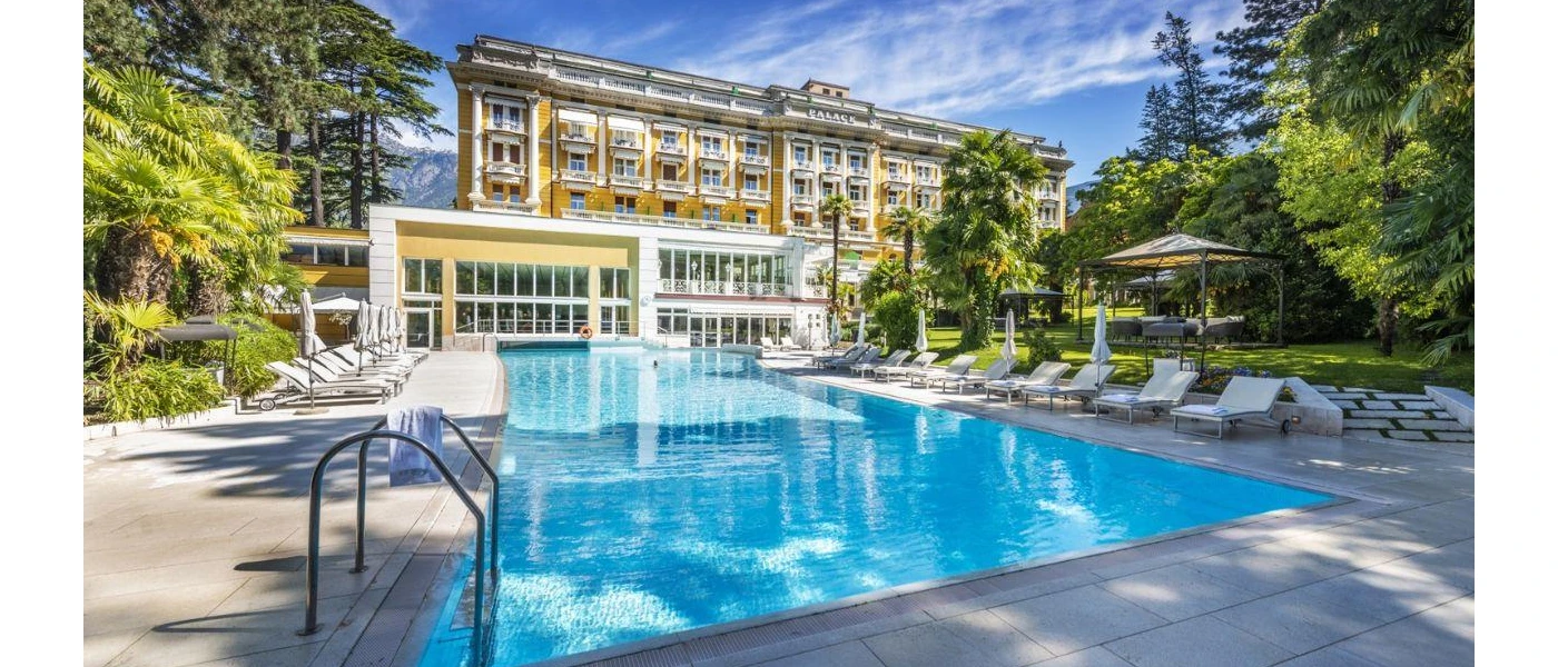 Grand multi-storey building with gold-yellow walls, overlooking a swimming pool surrounded by loungers and leafy greenery under a blue sky