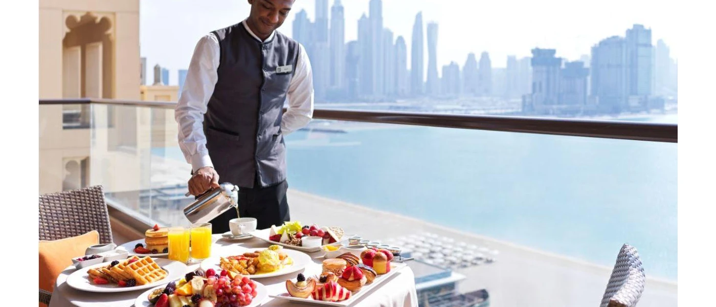 Smiling waiter pours coffee on a balcony with a view of the Dubai skyline, and a table laid with fruit, waffles and juice for breakfast