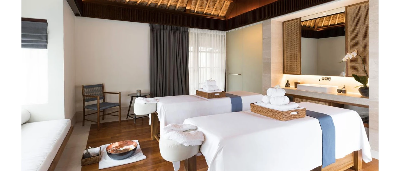 Spa treatment room with two beds covered in white, with rattan trays of white towels on top, and a vanity area with a potted lotus flower