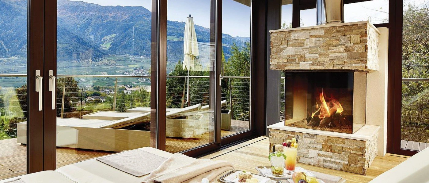 Hotel room with a stone fireplace, dining table and floor-to-ceiling balcony doors showcasing a terrace with loungers and views of the mountains