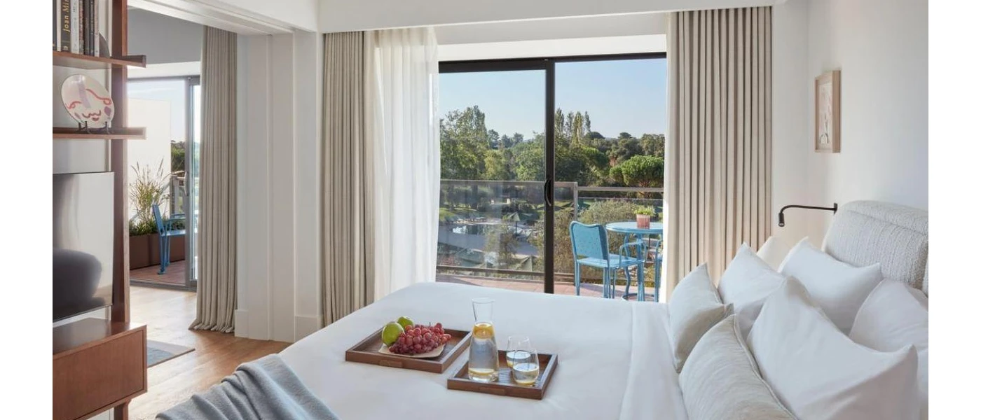 Hotel room with white walls and bedding, cream drapes, black-framed full-length balcony doors and a wooden cabinet 