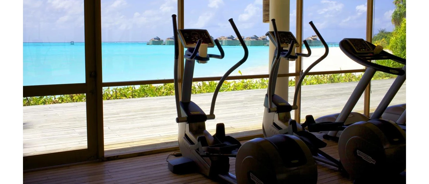 Bike machines in a gym with floor-to-ceiling windows overlooking white sands and turquoise shallows