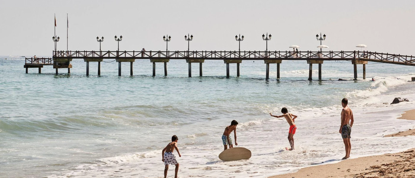 Man and three boys in swimwear play with a surfboard on the shoreline, with a wooden pier in the background