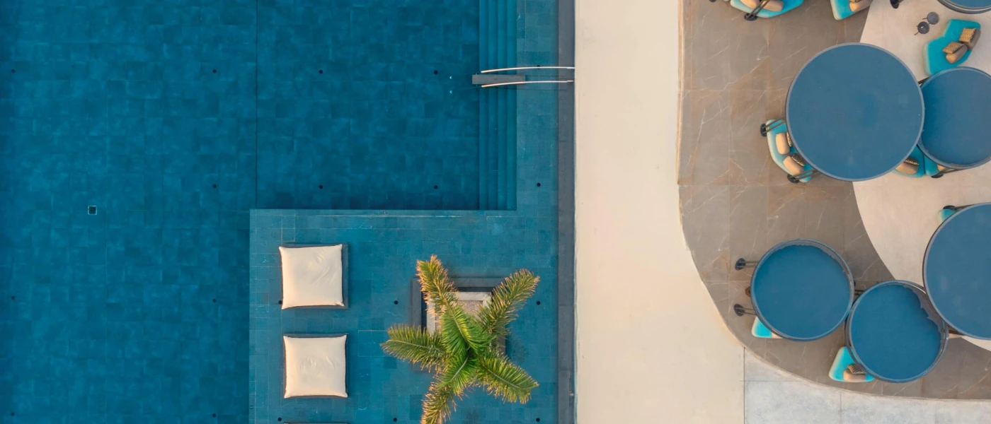 Tiled swimming pool with a palm island and giant cream cushions, next to a terrace with black pergolas and blue chairs