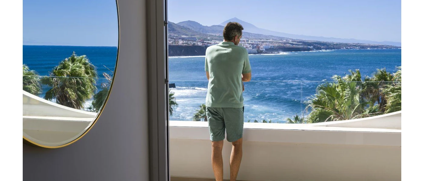 Man in shorts and t shirt looking out at the ocean and mountains from a balcony