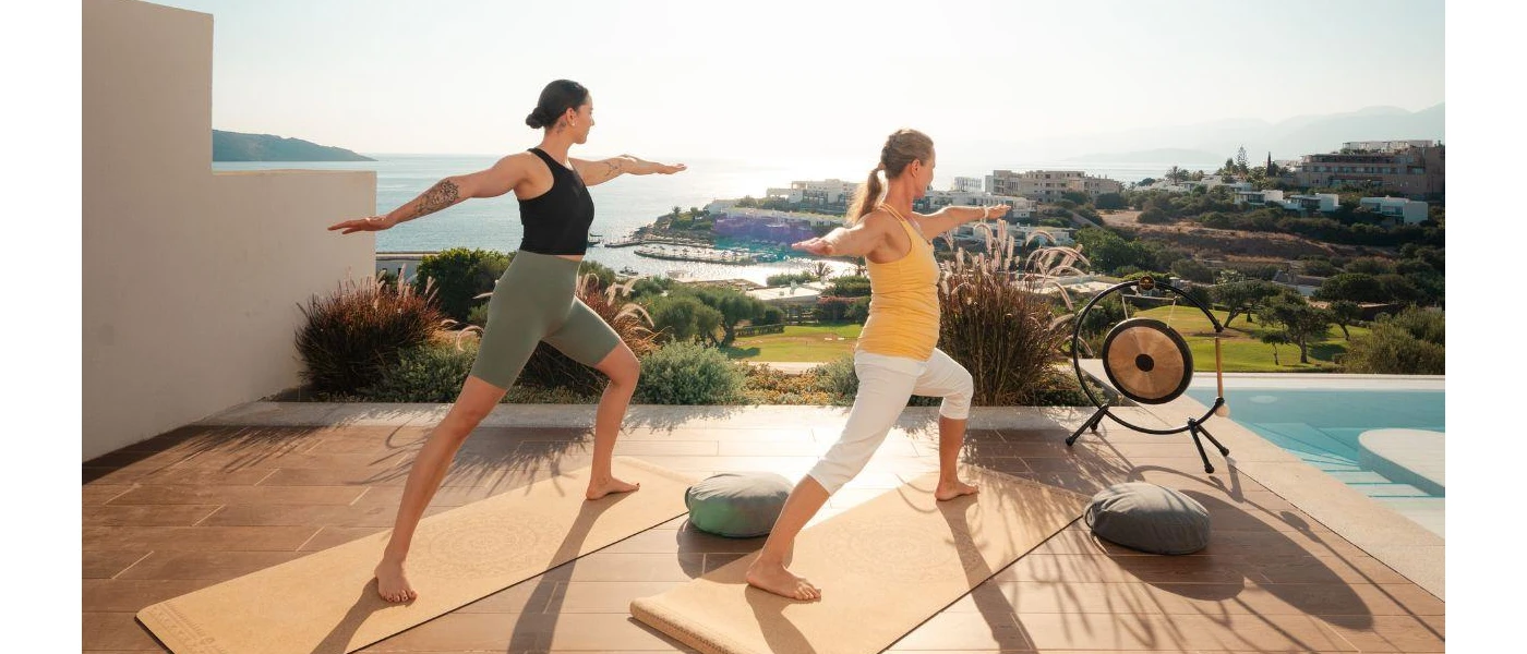 Two women in active wear in warrior pose on a wooden deck next to the pool and a large sound gong