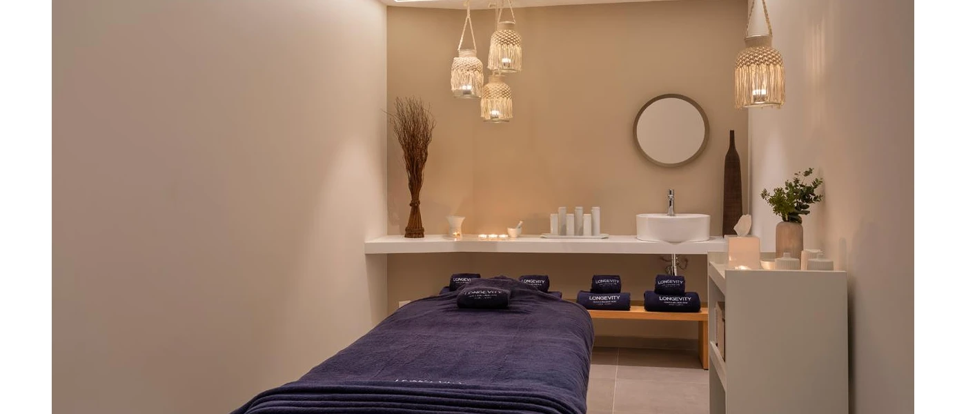 Spa treatment room with cream walls, a massage bed with blue towels, bohemian-style rattan pendant lights and vanity units with flowers and a vase, and a wall-hung mirror