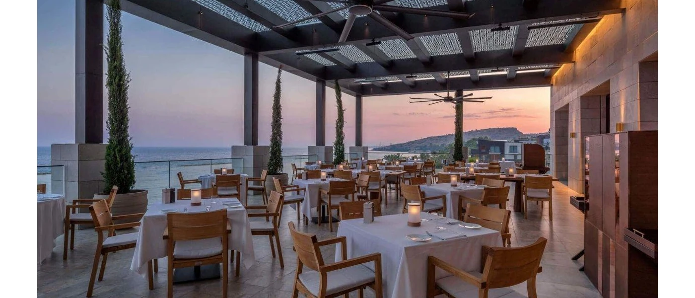 Outdoor restaurant terrace at sunset with tables laid out with crisp white tablecloths and an ocean view