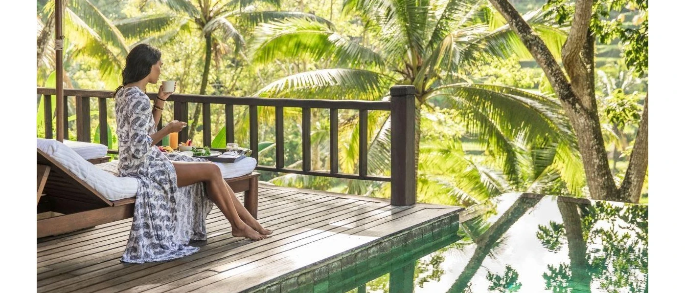Woman sipping from a white cup with a tray of food in front of her, sitting on a lounger and looking out over a tropical jungle