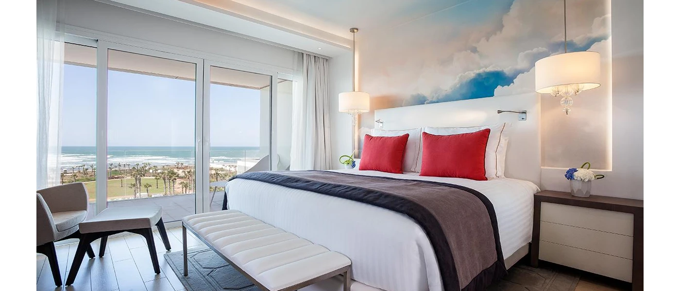 Hotel room with white bedding, red cushions and blue wallpaper and a large window