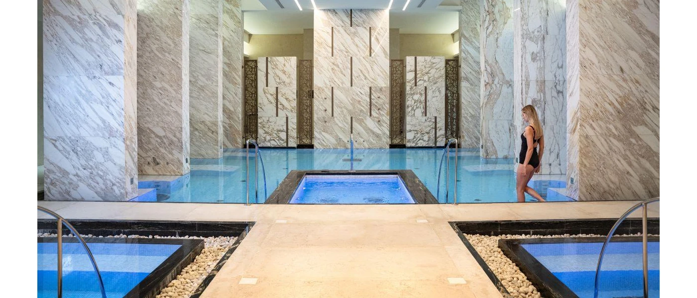 Indoor swimming pool and plunge pools surrounded by marble pillars, and a woman in a black swimsuit making her way into the water