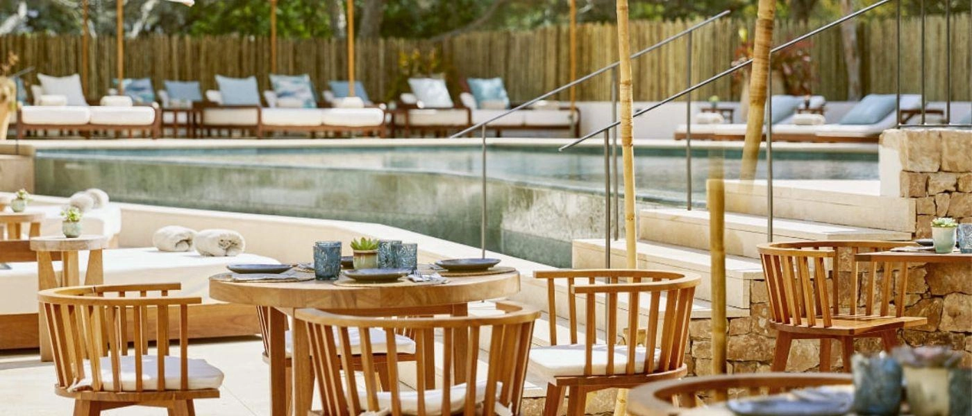 Swimming pool surrounded by wooden fencing, pine trees, white-cushioned loungers and wooden seating