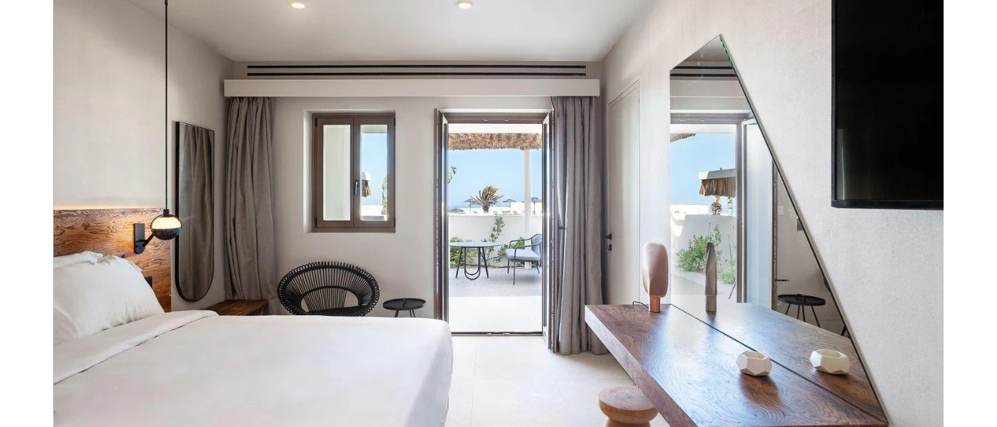 Chic hotel bedroom with white bedding, a wooden headboard, dressing table and large mirror, black iron-style chair and balcony with a table and chairs  