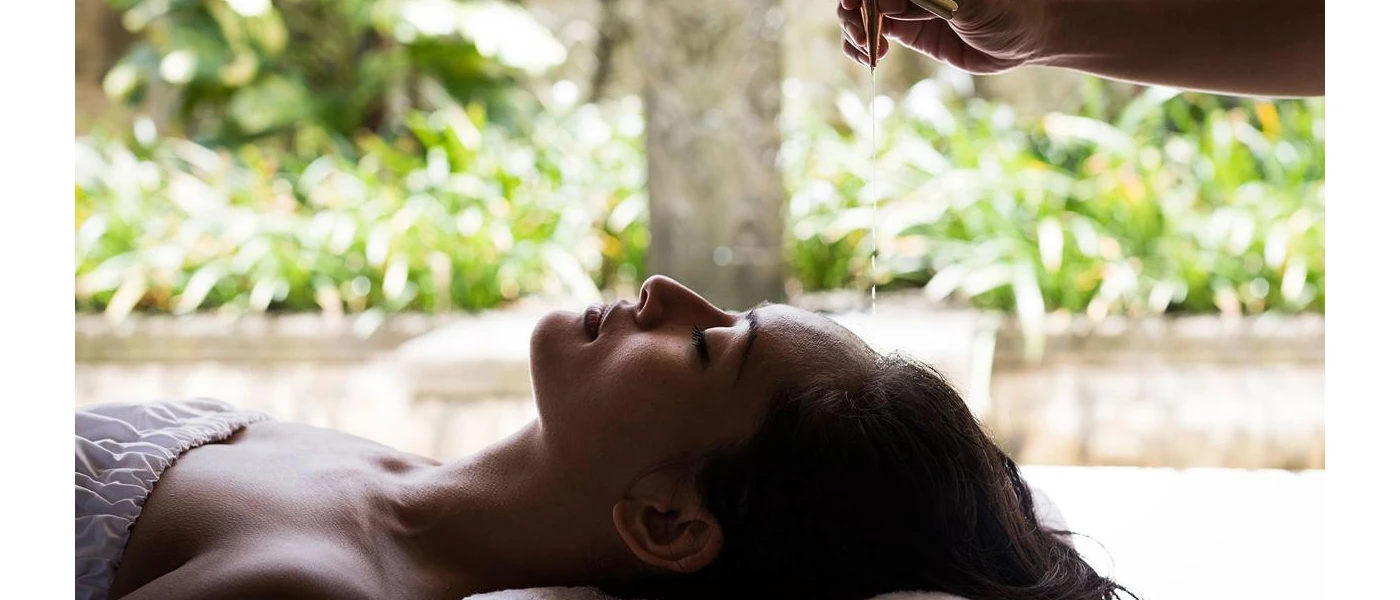 Woman with dark hair and closed eyes lying down surrounded by tropical greenery as a therapist pours oil on her head