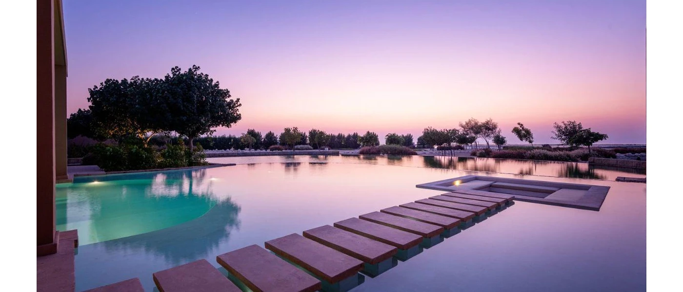 The sun sets over a swimming pool with wooden stepped walkways, sunken seating areas and a fringe of greenery