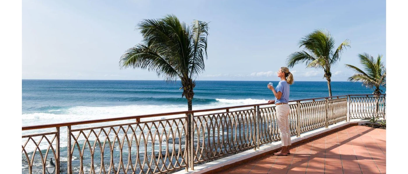 Woman in a blue t shirt looking out at palm trees and the ocean on a sunny paved terrace