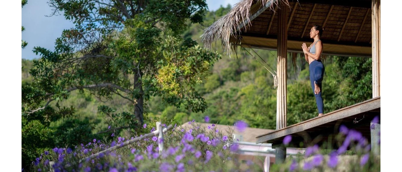 Woman stands in active wear at the edge of a thatched-roof open-sided pavilion with her hands clasped in prayer overlooking tropical greenery