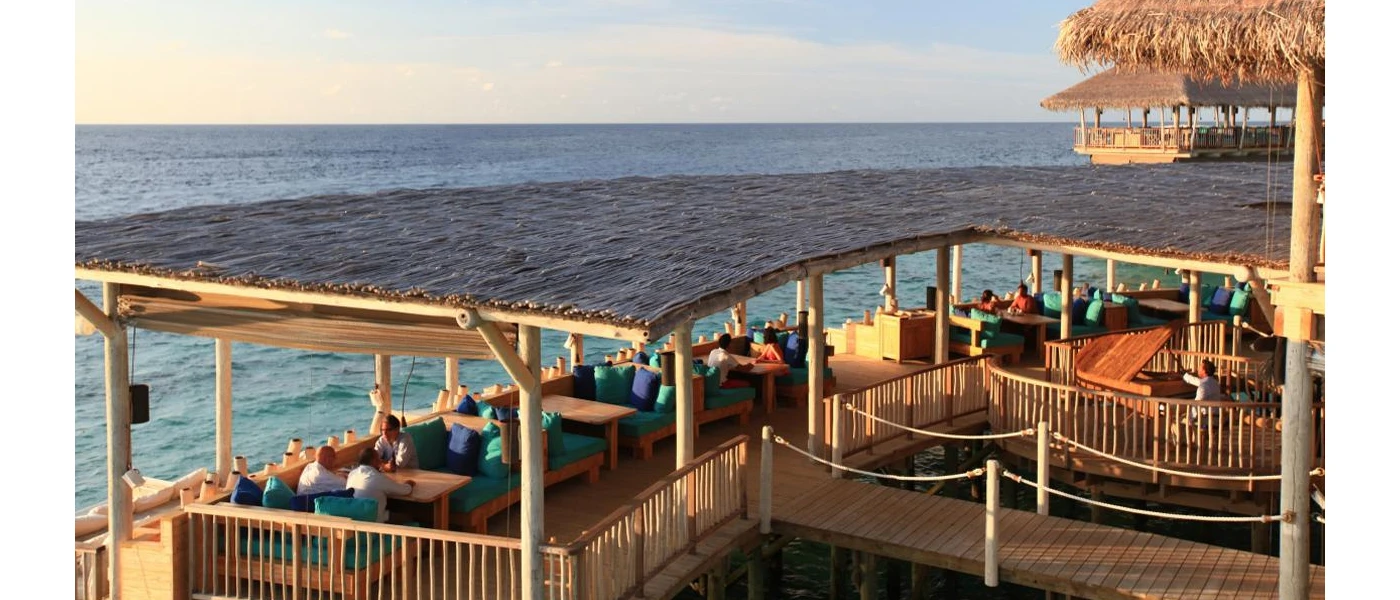 Thatched-roof building overlooking the ocean, with wooden tables and chairs. blue cushions, and a pianist playing a wooden piano