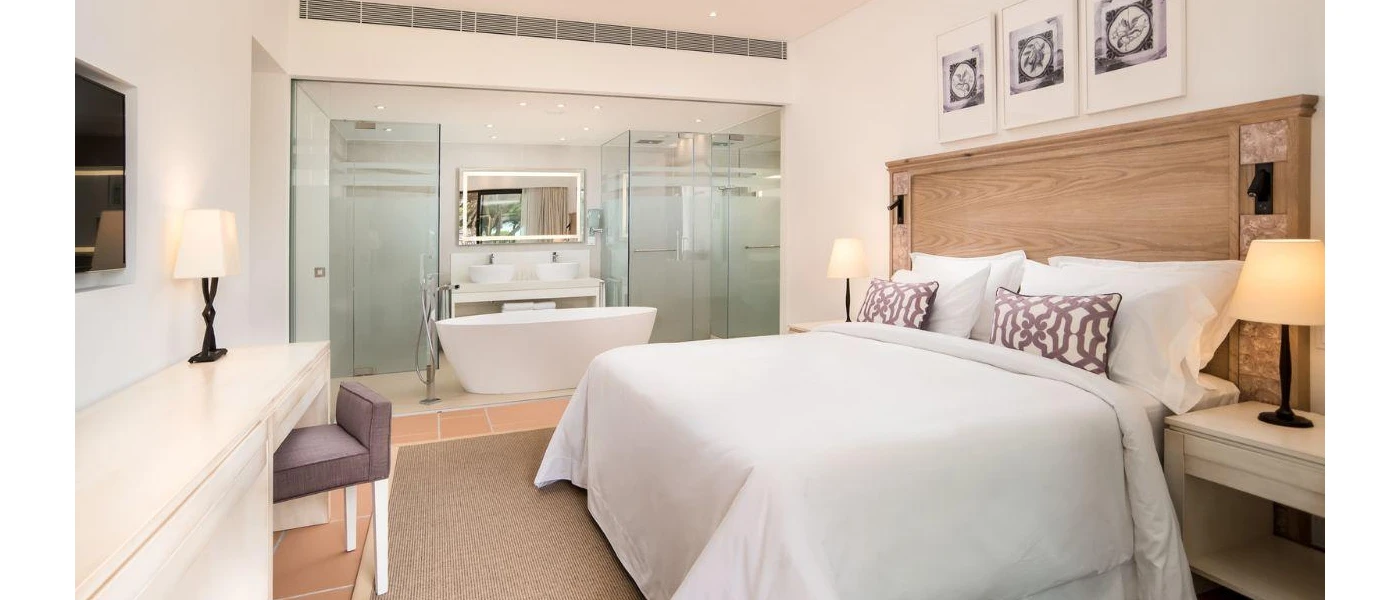 Hotel room with gleaming white walls, bedding and a standalone bathtub, a white desk, wall-hung TV and cream rug, along with a wooden headboard and black and white framed pictures above the bed