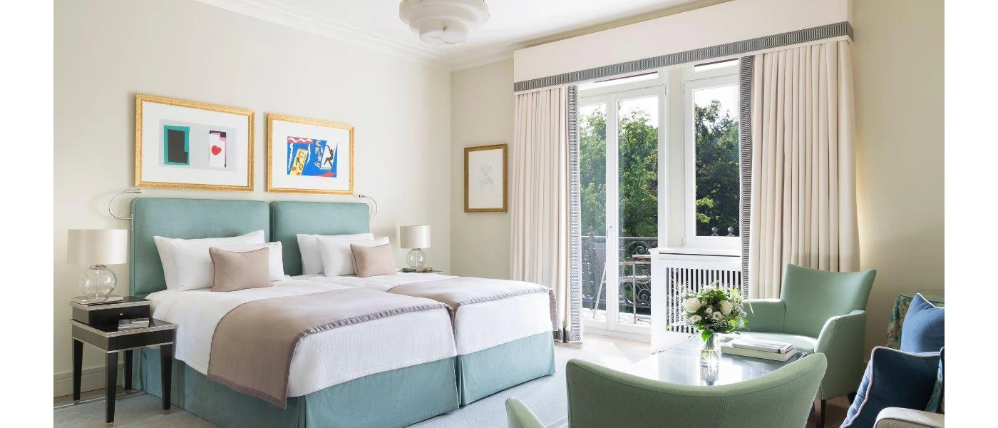 Stylish neutral hotel room with twin beds, curtains, teal armchairs and a vase of flowers, and contemporary art hung above the bed