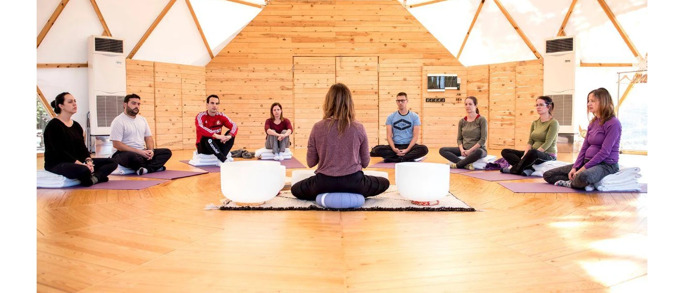 Group sits in a circle in a sheer domed building, while a woman sits at the front of the class next to two white sound healing bowls