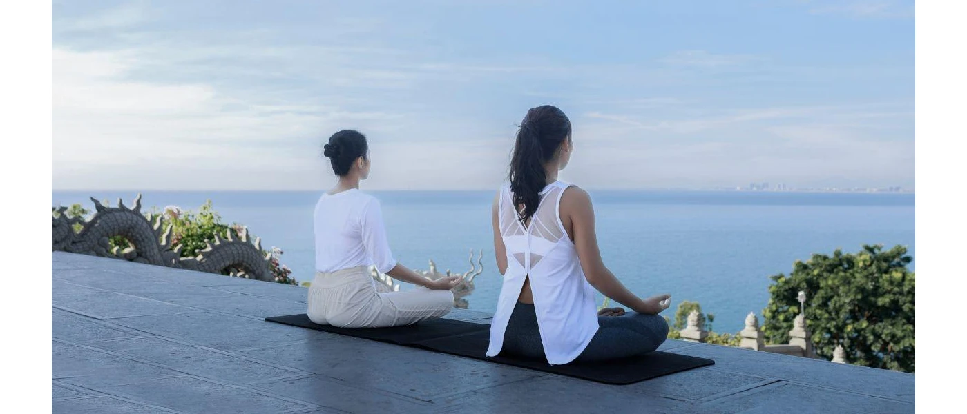 Two women sitting in lotus position on a yoga mat, facing the sea and tropical greenery below