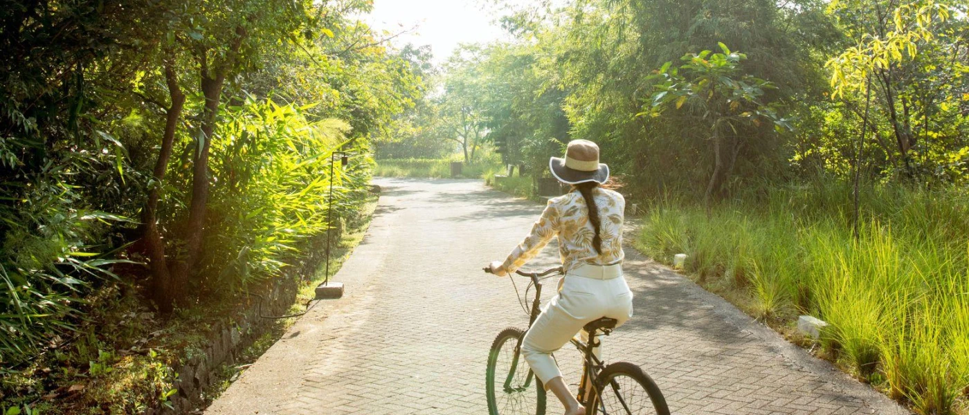 Woman in a hat, white trousers and a yellow floral patterned blouse cycles through a walkway surrounded by greenery