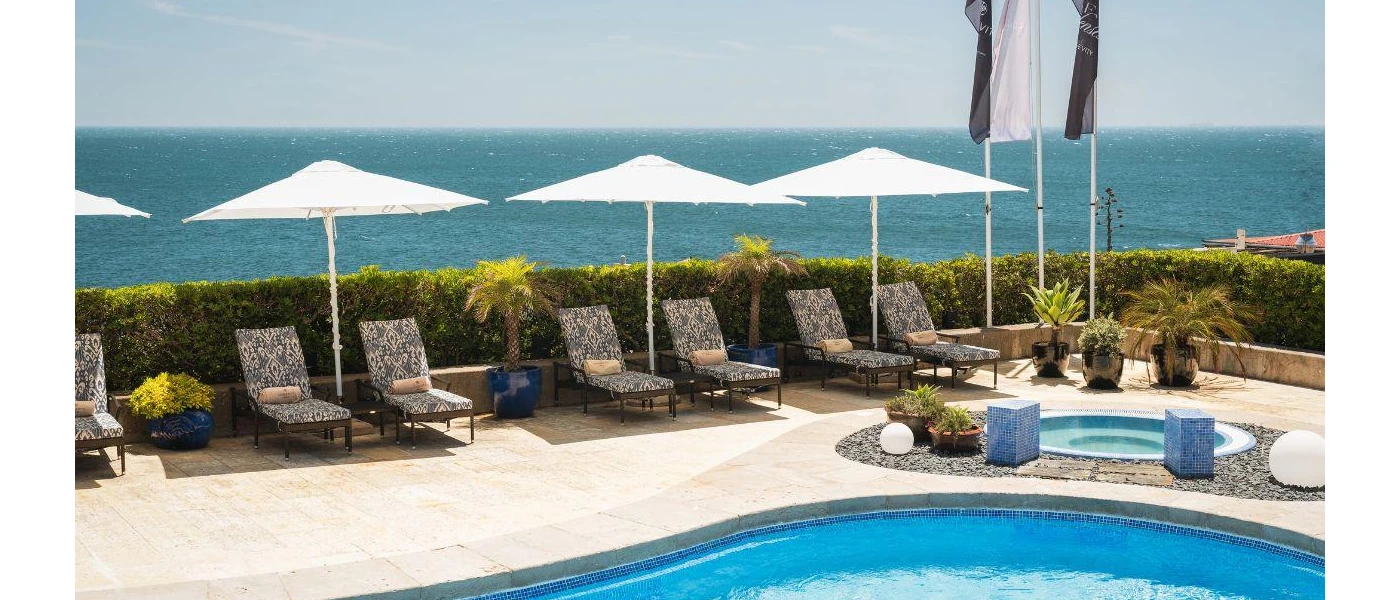 Curved swimming pool in a sunny terrace, with white parasols, loungers with blue and white floral cushions, a corner Jacuzzi and views of the ocean