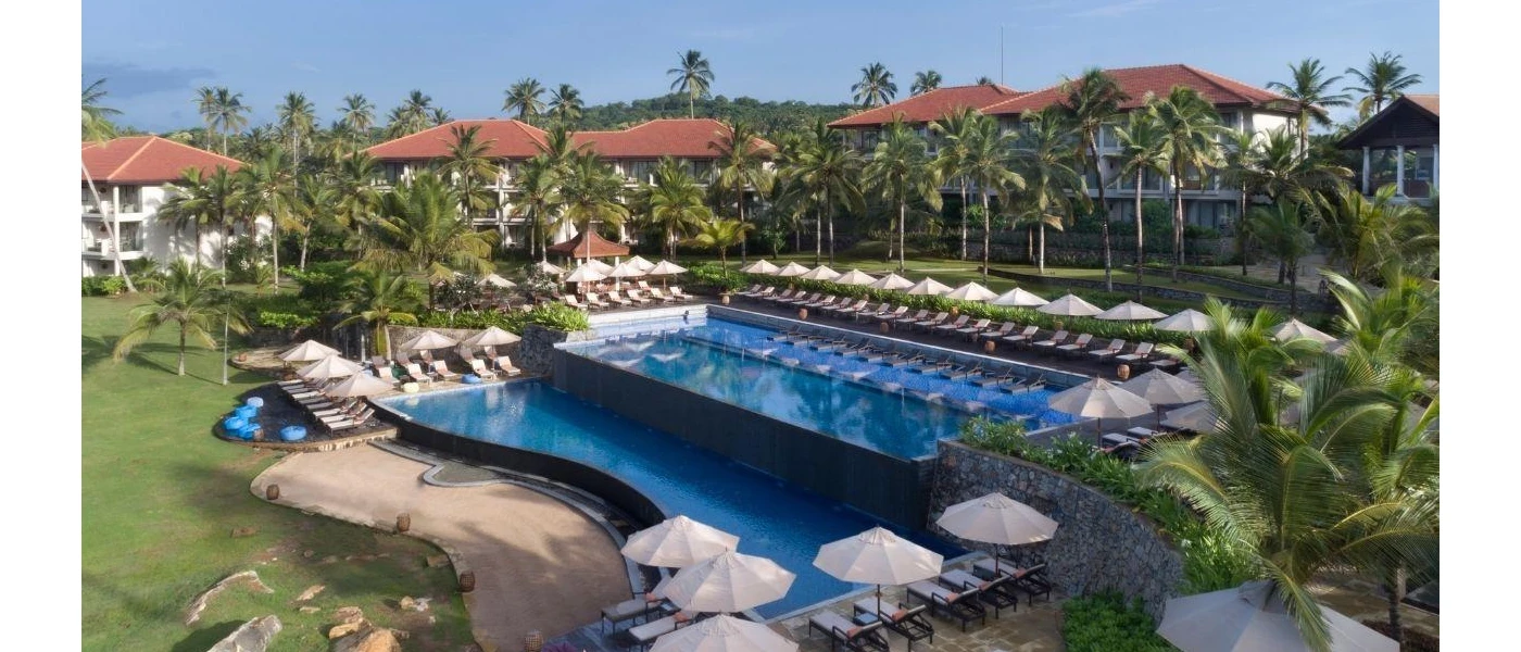 Split-level pool surrounded by white loungers and parasols, terracotta-roofed buildings and gardens filled with palm trees