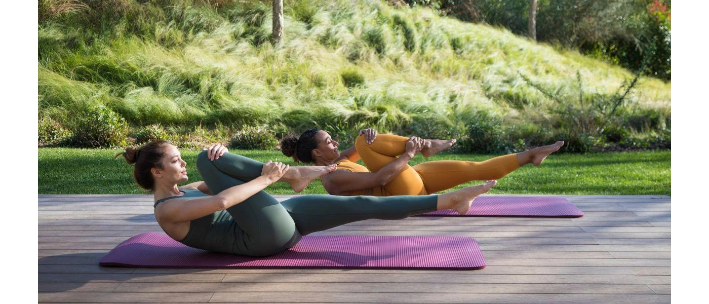 Two women in mustard and khaki gym sets on purple mats holding one knee into their chests