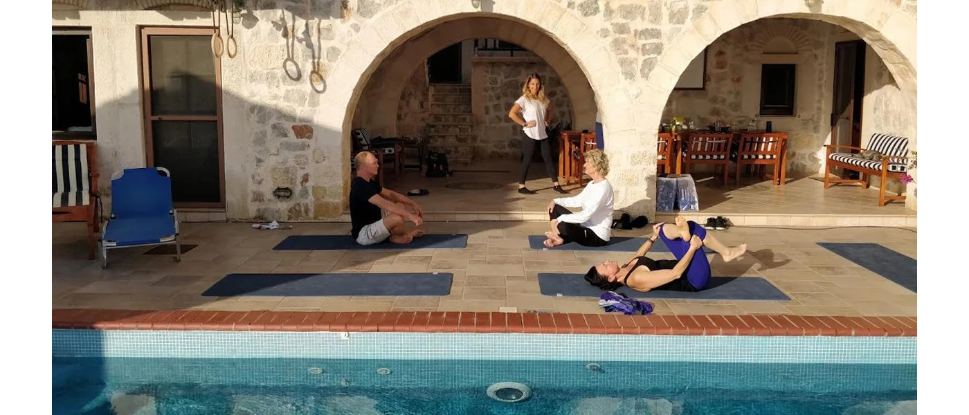 Three people stretching on blue yoga mats next to a pool, as an instructor watches on and the sun comes up