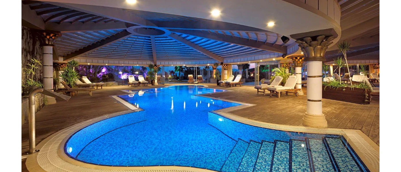 Indoor pool area with ceiling spotlights. white columns, tropical plants, white-cushioned loungers and floor-to-ceiling windows