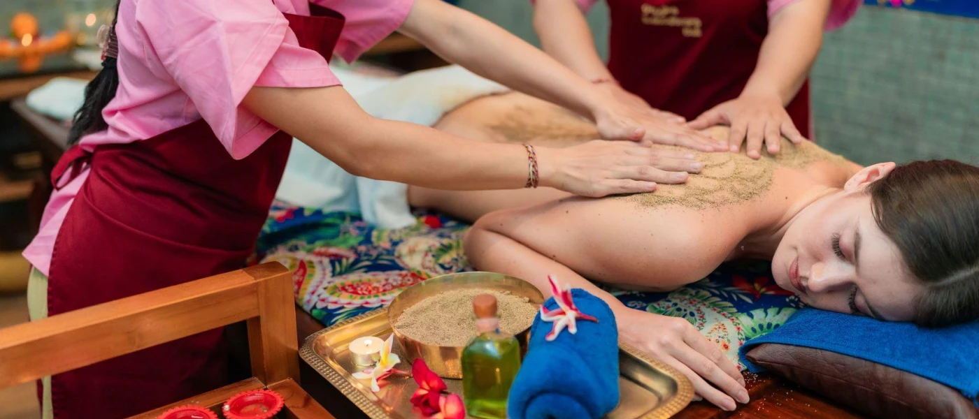 Woman with her eyes closed lying on a bed as two therapists in red aprons massage her back with a sand-like substance