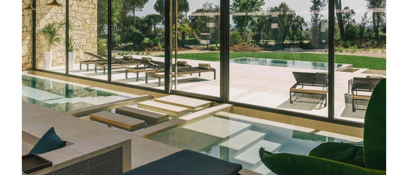 Indoor pool with stepped walkways and entry points, in front of floor-to-ceiling windows overlooking an outdoor pool